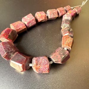 Peut inclure: Un fil de perles de pierres précieuses brun-rouge à facettes. Les perles sont de forme irrégulière, avec un mélange de tons foncés et clairs. Les perles sont enfilées sur un fil fin de couleur claire, créant un collier ou un bracelet.