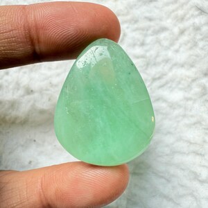 May include: A polished, teardrop-shaped gemstone in a light green hue. The stone has a smooth, translucent surface with subtle internal patterns. The gemstone is held between fingers, showcasing its size and shape.