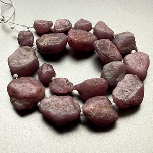 Può includere: Una collana di pietre preziose di rubino rosso-rosato grezze. Le pietre variano per dimensioni e forma, infilate con un sottile cordoncino bianco. Le pietre preziose hanno un aspetto naturale, non lucidato, con trame e variazioni di colore visibili.