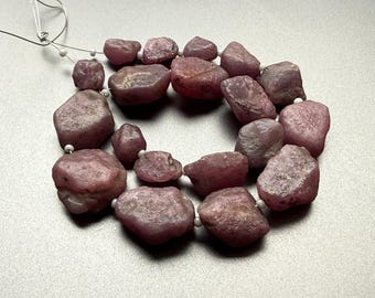 May include: A necklace of rough-cut, pinkish-red ruby gemstones. The stones vary in size and shape, strung together with a thin, white cord. The gemstones have a natural, unpolished appearance, with visible textures and variations in color.
