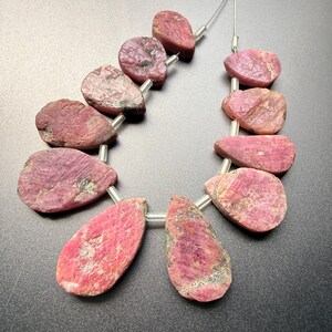 Könnte beinhalten: Eine Kette aus rohen Rubin-Edelsteinen. Die Steine haben eine tiefrosa-rote Farbe mit natürlichen Texturen und Variationen. Jeder Stein ist unregelmäßig geformt und mit einer transparenten Schnur aufgereiht.