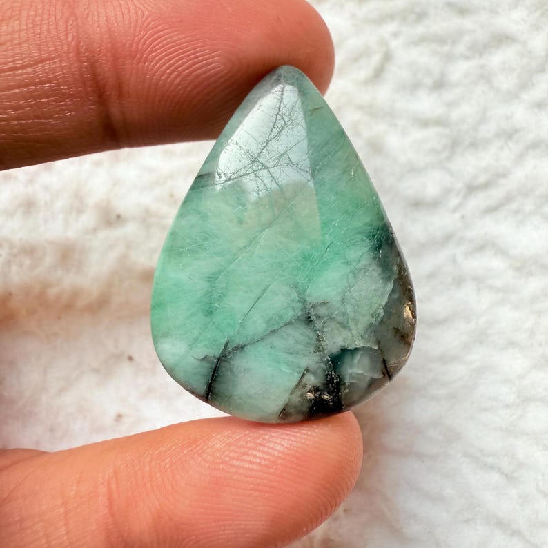 May include: A pear-shaped, faceted emerald gemstone. The gem features a vibrant green hue with dark inclusions and a polished surface. The stone is held between fingers, showcasing its size and clarity. The gemstone is a natural, untreated emerald.