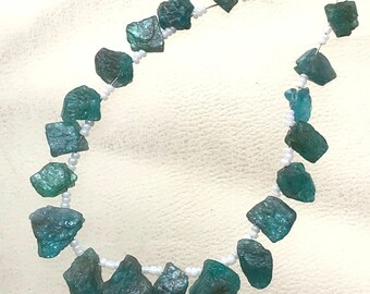 Puede incluir: Un collar con piedras preciosas de color turquesa en bruto, ensartadas en un hilo de cuentas blancas. Las piedras tienen una forma irregular y diferentes tonos de turquesa. El collar se muestra sobre un fondo blanco.