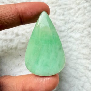 May include: A teardrop-shaped, polished gemstone in shades of light green. The stone has a smooth, glossy surface and is held between two fingers. The gemstone's color is consistent throughout, with subtle variations in tone.