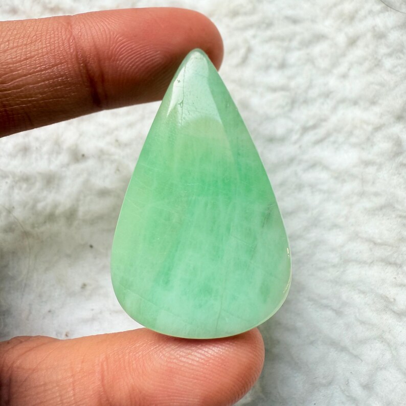 May include: A teardrop-shaped, polished gemstone in shades of light green. The stone has a smooth, glossy surface and is held between two fingers. The gemstone's color is consistent throughout, with subtle variations in tone.