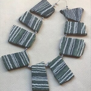 May include: A necklace made of green and white striped stone beads. The beads are rectangular and have a natural, rough texture. The necklace is strung on a silver chain.