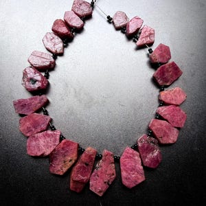 Peut inclure: Un collier composé de pierres précieuses de rubis brutes et irrégulières dans des tons rose foncé et magenta. Les pierres sont enfilées avec de petites perles d'espacement noires rondes sur un fil fin et flexible, formant un cercle.