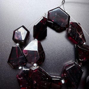 May include: A strand of deep red, faceted garnet beads with irregular, geometric shapes. The translucent, dark burgundy stones are strung on a thin silver-toned wire, separated by small white spacer beads, showcasing natural inclusions and reflective surfaces.