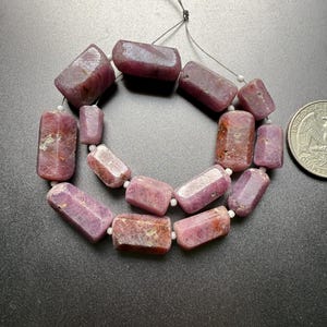 May include: A necklace of faceted, rectangular ruby beads in shades of pink and red. The beads are strung together with a thin, dark string and small white beads. A U.S. quarter is in the image for size comparison.