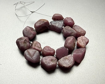May include: A strand of rough-cut, tumbled gemstones in shades of pink and purple. The stones are strung together with a thin, white cord, forming a circular shape. The gemstones have a natural, unpolished appearance.