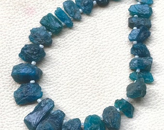 Puede incluir: Un collar de piedras preciosas en bruto de color azul verdoso. Las piedras varían en tonalidad, desde el verde azulado oscuro hasta el turquesa claro, ensartadas con pequeñas cuentas blancas. Las formas irregulares de las piedras dan al collar un aspecto natural y orgánico.