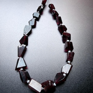 May include: A strand of deep red, irregularly shaped garnet beads arranged in a teardrop loop. Each dark, translucent gemstone is separated by a small, round white spacer bead, all strung together on a thin, flexible silver-toned wire.