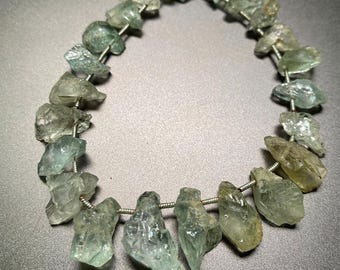 May include: A strand of light green aquamarine gemstone beads strung on a silver wire. The beads are irregular in shape and have a natural, rough texture.