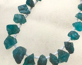 Puede incluir: Un collar con piedras preciosas de color turquesa en bruto ensartadas en un hilo de cuentas blancas. Las piedras tienen una forma natural e irregular, sobre un fondo blanco.