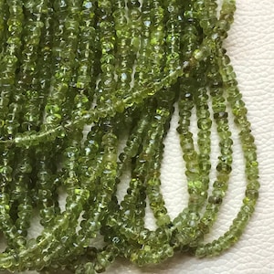 Op de afbeelding: Een streng groene peridot kralen, gefacetteerd en rond, met een gladde, gepolijste afwerking. De kralen zijn geregen op een doorzichtige draad en zijn klaar om te worden gebruikt voor het maken van sieraden.