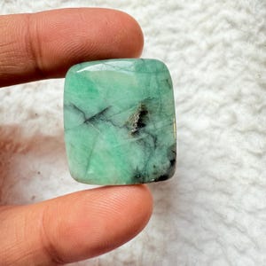 May include: A square-cut, polished emerald gemstone. The gem is a vibrant green color with dark inclusions and a slightly rounded shape. The emerald is held between two fingers, showcasing its natural beauty and clarity.