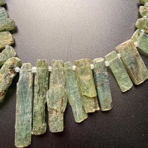 May include: A necklace strand featuring raw, elongated, light green kyanite crystal segments. Each rectangular stone displays a natural, textured surface with visible striations and inclusions, separated by small, clear, round spacer beads.