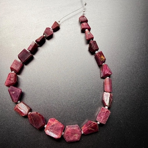May include: A strand of raw, deep pinkish-red ruby gemstone beads arranged in a U-shape. The beads are irregularly shaped, faceted, and vary in size, separated by small white spacers along a thin, clear filament wire for jewellery making.