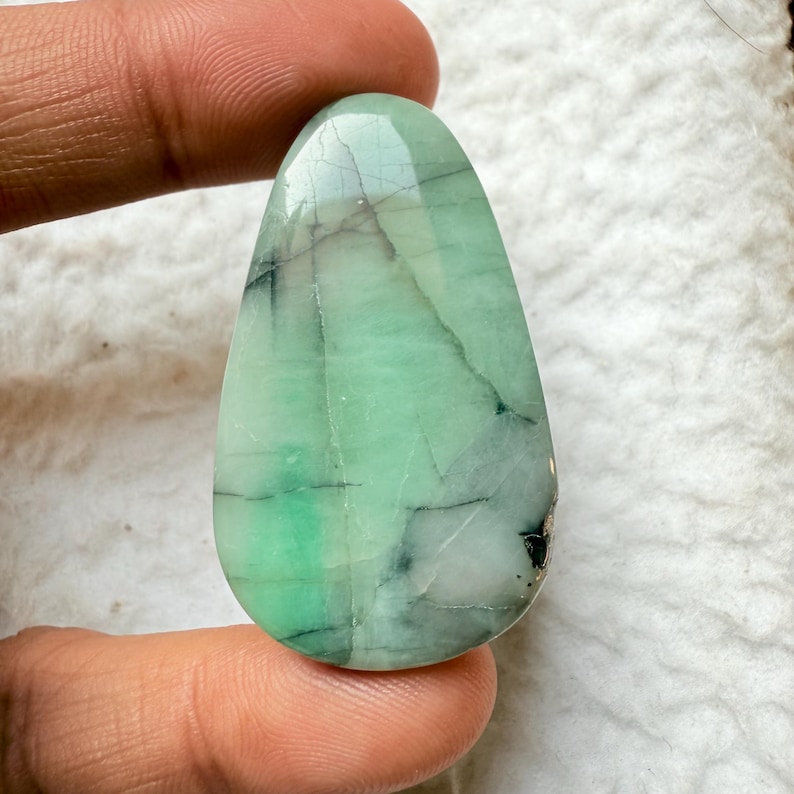 May include: A polished, teardrop-shaped gemstone with a light green hue, veined with dark gray and black inclusions. The stone has a smooth, reflective surface, and is held between fingers.