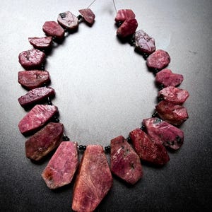 Pode incluir: Um colar com uma série de pedras de rubi brutas de cor rosa profundo dispostas num padrão graduado. As pedras são separadas por pequenas contas pretas redondas e enfiadas num fio fino, criando uma forma circular com um acabamento natural.