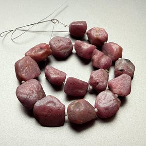 Puede incluir: Un collar de piedras preciosas en bruto, de color rosa rojizo. Las piedras tienen forma irregular y están ensartadas con un fino alambre plateado. Pequeñas cuentas blancas separan cada piedra. El fondo es de color claro y neutro.