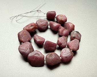 May include: A necklace of rough-cut, reddish-pink gemstones. The stones are irregularly shaped and strung together with a thin, silver-colored wire. Small white beads separate each stone. The background is a light, neutral color.