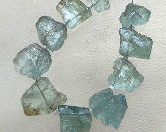 May include: A strand of rough-cut, pale blue-green aquamarine gemstone beads. The irregular, translucent stones are strung together with a thin, clear wire and small white beads. The beads have a natural, raw appearance.