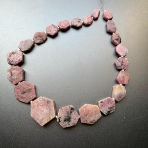 以下が含まれることがあります： 暗いインクルージョンを持つ、ファセット加工されたピンク色の宝石のネックレス。石は細い白い糸で繋がれています。ネックレスは濃い灰色の表面に表示されています。