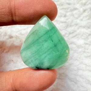 May include: A teardrop-shaped, polished gemstone in shades of green. The gem has a smooth, reflective surface with visible internal striations. The stone is held between two fingers, showcasing its size and shape. The gem is likely used for jewelry.