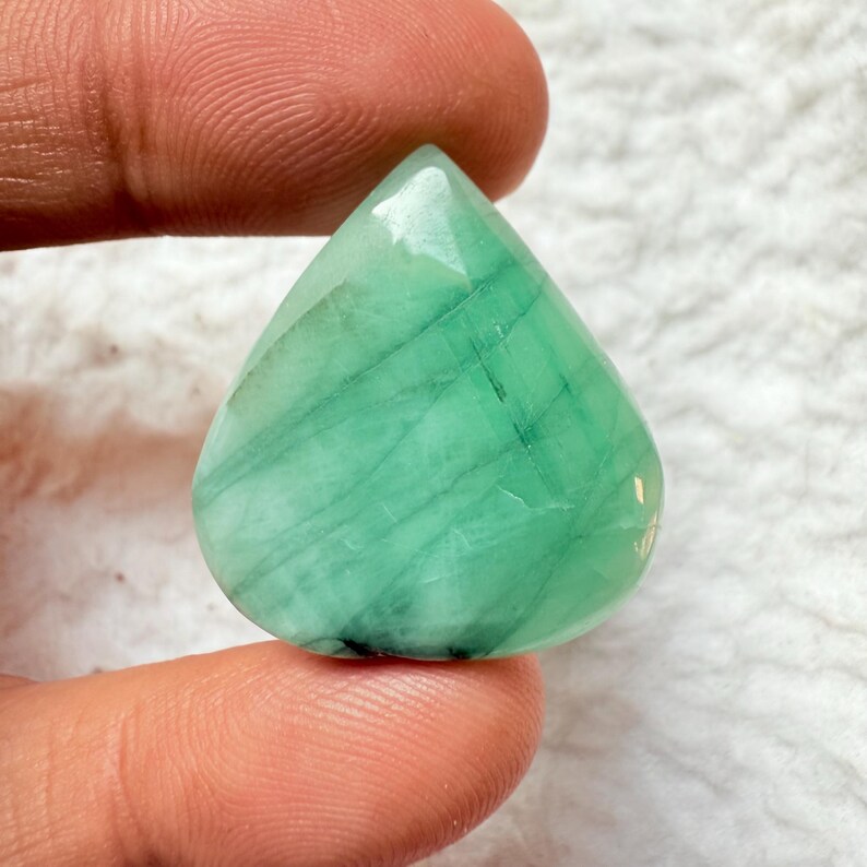 May include: A teardrop-shaped, polished gemstone in shades of green. The gem has a smooth, reflective surface with visible internal striations. The stone is held between two fingers, showcasing its size and shape. The gem is likely used for jewelry.