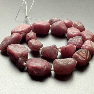May include: A necklace of rough-cut, pinkish-red ruby beads. The beads are irregular in shape and size, strung together with white string. The gemstones have a natural, unpolished appearance, with variations in colour and texture.