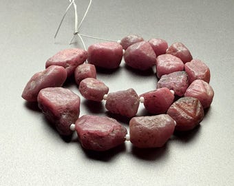 May include: A strand of rough-cut, pinkish-red ruby beads. The beads are irregular in shape and size, strung together with white string. The gemstones have a natural, unpolished appearance, with variations in color and texture.