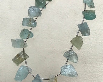 May include: A strand of rough-cut aquamarine gemstones in varying shades of blue and green. The stones are strung on a thin, silver-colored wire, creating a necklace-like arrangement. The gemstones have a natural, irregular shape.