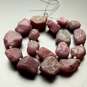 Peut inclure: Un collier de pierres précieuses roses brutes. Les pierres, de tailles et de formes variées, sont enfilées sur un fil fin avec de petites perles blanches. Les pierres ont un aspect naturel et non poli, avec des variations de couleur et de texture.
