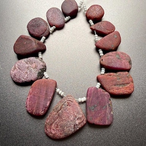 Peut inclure: Un collier composé de pierres précieuses rouge-violet foncé de forme irrégulière. Les pierres sont enfilées sur un fin cordon de perles blanches. Les pierres ont une texture rugueuse et naturelle. Un bijou unique.
