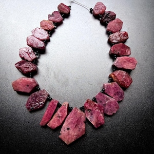 May include: A necklace featuring a series of raw, irregularly shaped deep pink ruby stones strung together with small, round black spacer beads. The stones vary in size and texture, arranged in a graduated pattern with the largest piece at the centre.