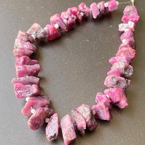 May include: A strand of pink ruby gemstone beads, each bead is a rough, irregular shape. The beads are strung on a white thread.