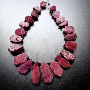 Puede incluir: Un collar compuesto por piedras de rubí en bruto de color rosa intenso con formas irregulares, dispuestas en forma de lágrima. Las piedras están separadas por pequeñas cuentas negras redondas ensartadas en un hilo fino, creando un aspecto orgánico.