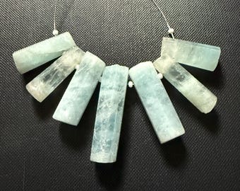 May include: A necklace featuring rectangular, light blue aquamarine gemstones. The stones are strung together with a thin, silver-colored wire and small white beads. The gemstones have a translucent quality and are arranged in a fan-like pattern.