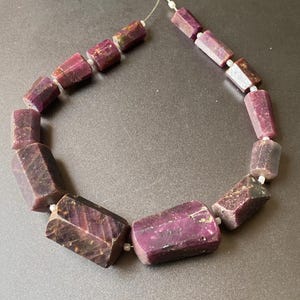 Puede incluir: Un collar de cuentas de piedras preciosas facetadas, de color rojo púrpura oscuro. Las cuentas son rectangulares con bordes angulados y están ensartadas en un fino alambre plateado. Las piedras preciosas tienen un aspecto natural y texturizado.