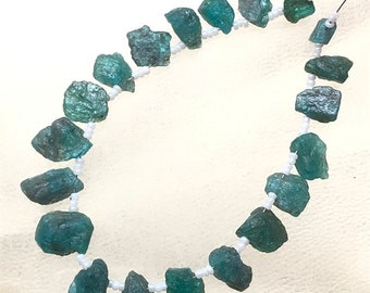 Puede incluir: Un collar con piedras preciosas en bruto de color verde azulado, ensartadas en un hilo de cuentas blancas. Las piedras preciosas tienen un aspecto natural y texturizado. El collar se muestra sobre un fondo blanco.