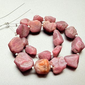 May include: A necklace of pink, irregularly shaped gemstones, possibly tourmaline or rhodochrosite, strung on a wire with small white beads. The stones vary in size and shade, with some showing an orange hue. The gemstones are approximately 1.3 to 2.5 cm in size.