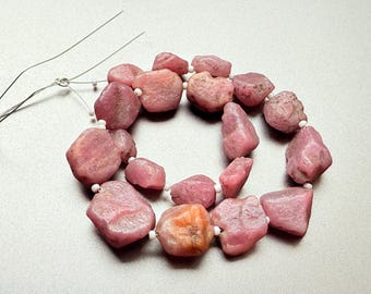 May include: A strand of pink, irregularly shaped gemstones, possibly tourmaline or rhodochrosite, strung on a wire with small white beads. The stones vary in size and shade, with some showing an orange hue. The gemstones are approximately 0.5 to 1 inch in size.