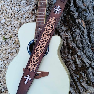 May include: A brown leather guitar strap with an intricate Celtic knot design and a silver cross. The strap is decorated with a brown and gold pattern. The strap is attached to a white acoustic guitar.