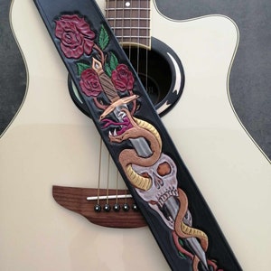 Może przedstawiać: Czarny skórzany pasek do gitary z motywem czaszki, węża, sztyletu i róż. Pasek jest na białej gitarze akustycznej.