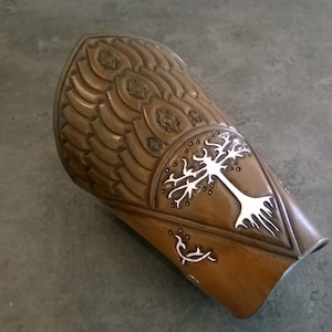 May include: A brown leather arm guard with a white tree design and a decorative pattern. The arm guard is made of leather and has a textured surface.