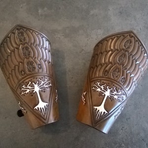 Puede incluir: Un par de protectores de brazo de cuero marrón con un diseño de árbol blanco. Los protectores tienen una superficie texturizada y están decorados con un patrón repetitivo.