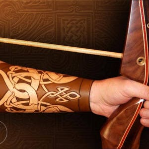 May include: A brown leather arm guard with an intricate Celtic knot design. The arm guard is worn on the left arm of a person holding a wooden bow.