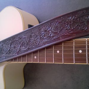Può includere: Un cinturino per chitarra in pelle marrone con un intricato disegno floreale. Il cinturino è appoggiato su una chitarra acustica marrone chiaro.