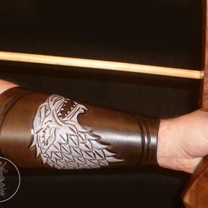 Peut inclure: Un protège-bras en cuir marron avec un motif de tête de loup argenté. La tête de loup a un design détaillé avec des dents acérées et une crinière texturée. Le protège-bras est porté sur le bras gauche d'une personne.